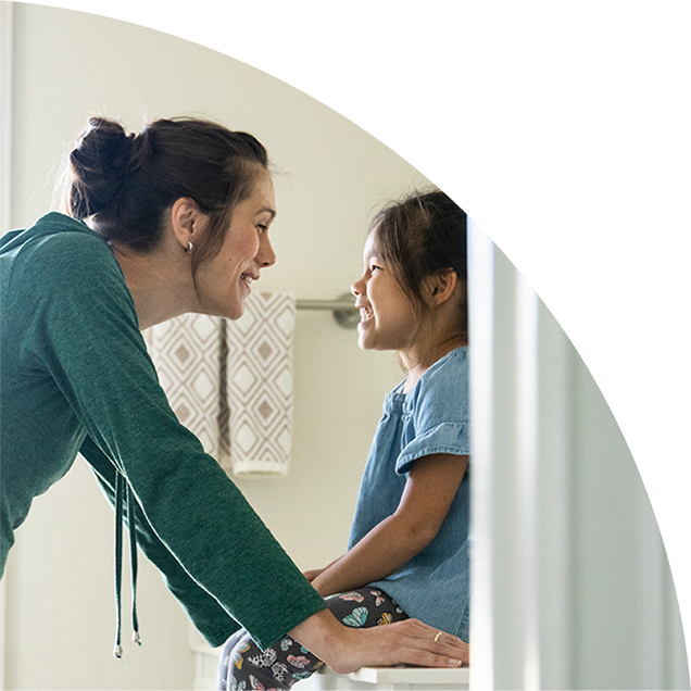 A kid with a big smile sitting on the bathroom vanity looks up at their watchful parent who's smiling back at them.